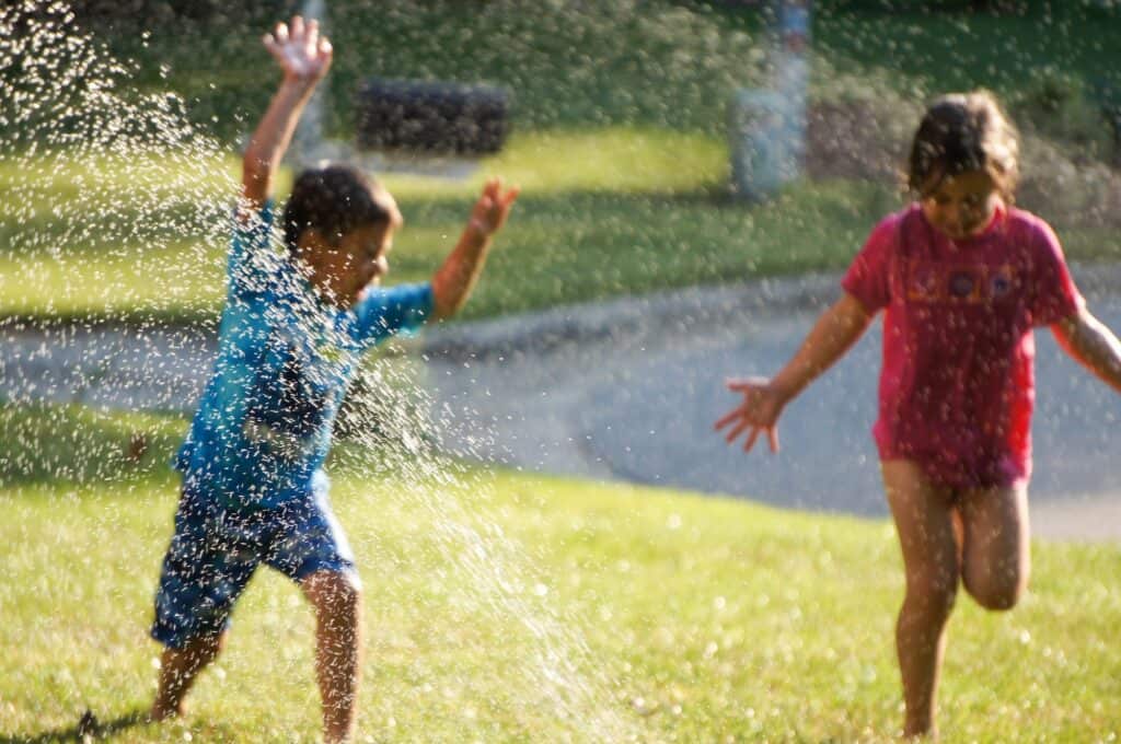Tips To Host Summer Celebrations at Home 2 Two children running through a sprinkler and playing outdoors in the summer
